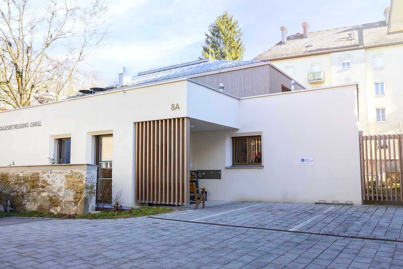 Flacher moderner Bau mit weißen Wänden, Holzlamellen und der Aufschrift Tagesbetreuung Gnigl.© SARA BUBNA photography