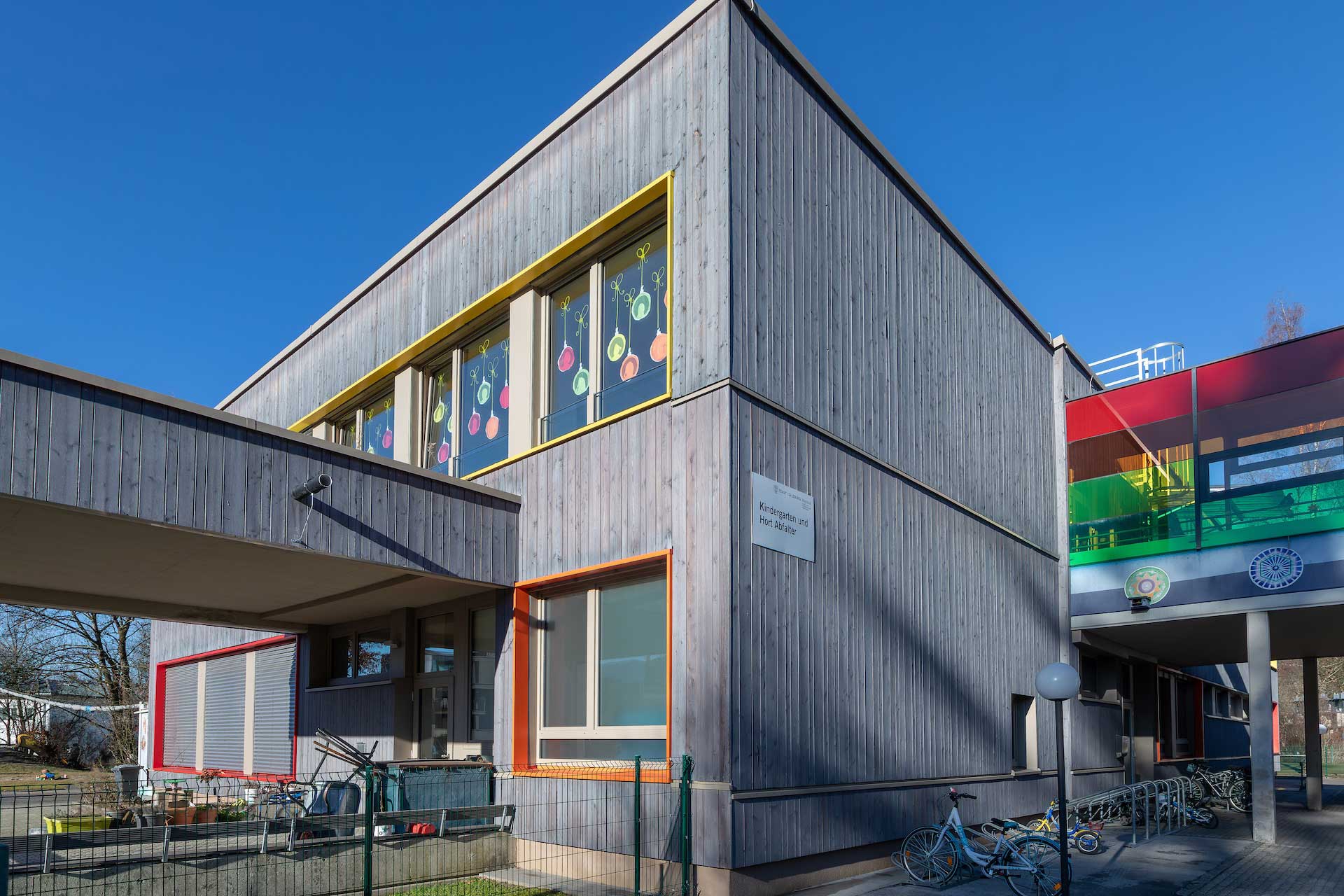 Außenansicht der neu mit Holzverkleidung und bunten Fensterrahmen sanierten Volksschule Abfalter an sonnigem Tag vor blauem Himmel, überdachter Eingangsbereich, bunte Fensterdekorationen © SARA BUBNA photography