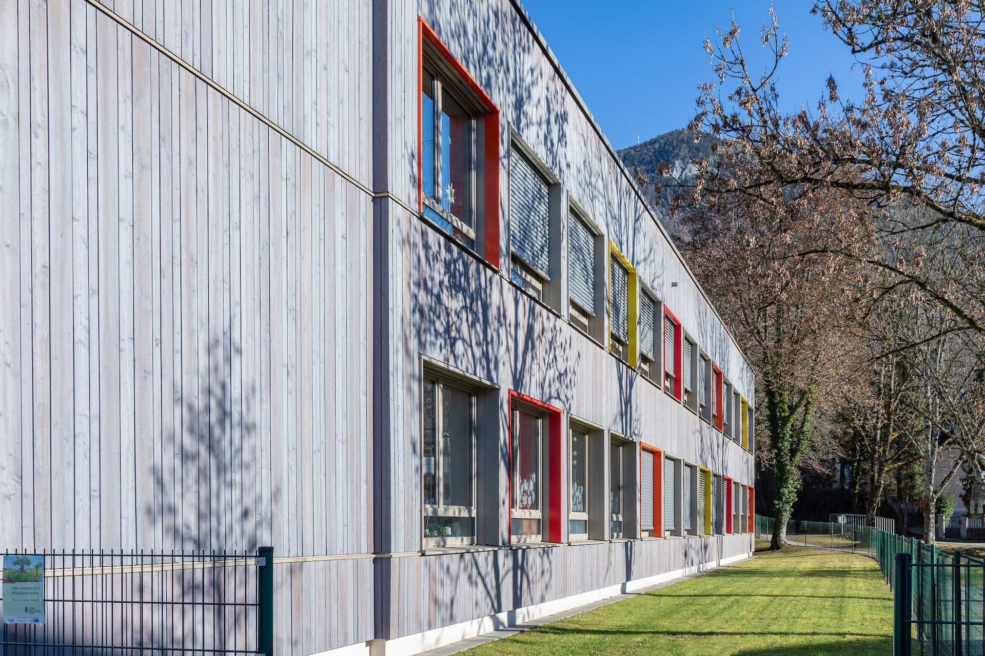 Außenansicht der neu mit Holzverkleidung und bunten Fensterrahmen sanierten Volksschule Abfalter an sonnigem Tag vor blauem Himmel, Garten mit altem Baumbestand, im Hintergrund schneebedeckter Gaisberg © SARA BUBNA photography