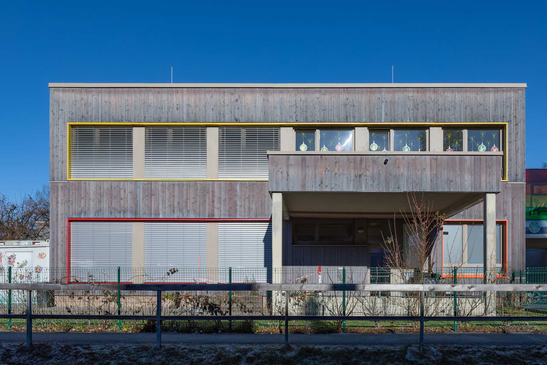 Außen-Frontalansicht der neu mit Holzverkleidung und bunten Fensterrahmen sanierten Volksschule Abfalter an sonnigem Tag vor blauem Himmel, Überdachter Eingang / Balkon, Fenster mit bunten Dekoelementen und tw. herabgelassenen Jalousien © SARA BUBNA photography