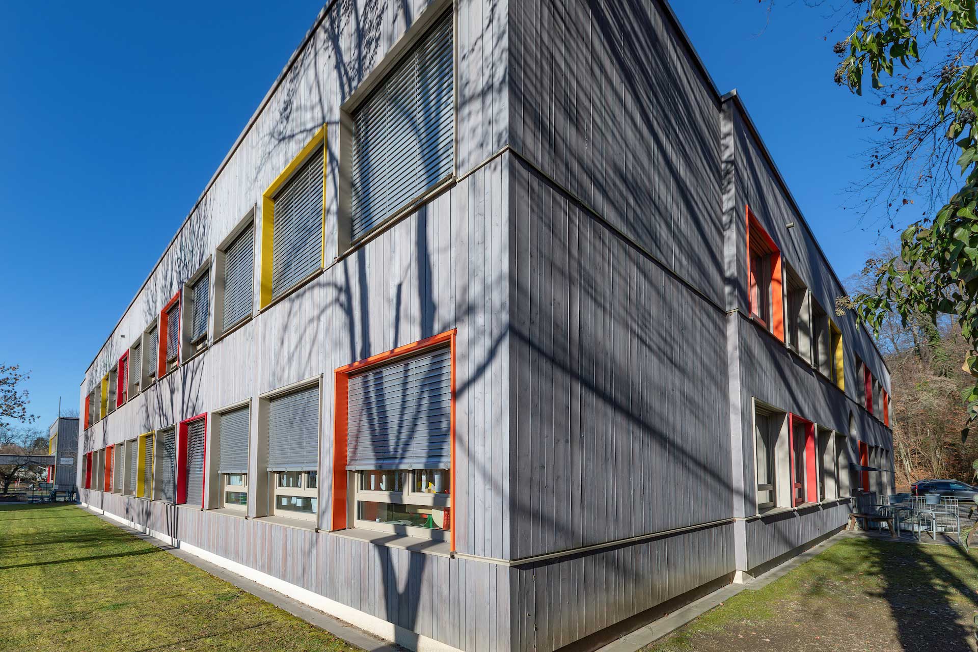 Außenansicht der neu mit Holzverkleidung und bunten Fensterrahmen sanierten Volksschule Abfalter an sonnigem Tag vor blauem Himmel © SARA BUBNA photography