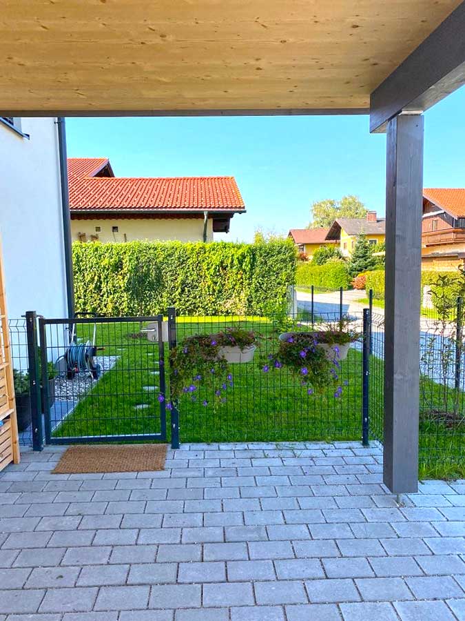„Kleiner Gartenbereich mit gepflasterter Terrasse, Holzüberdachung, Metallzaun, zwei Pflanztrögen mit violetten Blumen, Hecke und Blick auf Häuser mit roten Dächern.