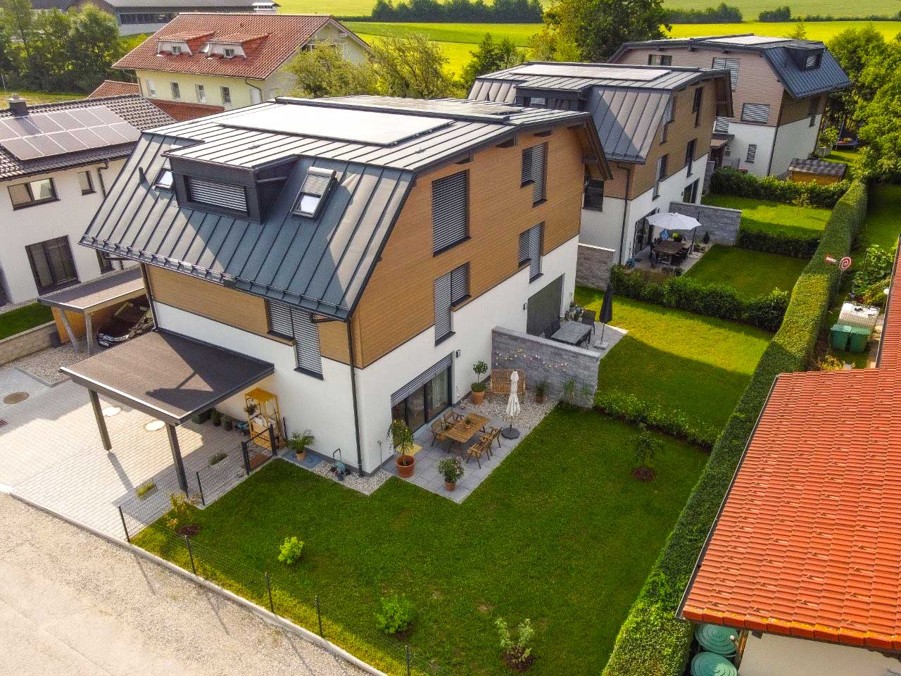 Moderne Wohnsiedlung mit mehrstöckigen Häusern, schrägen Metalldächern, Holzverkleidung, weißen Fassaden, kleinen Gärten und Sitzbereichen
