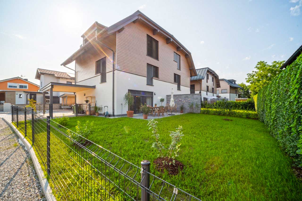 Modernes Wohnhaus mit hellem Putz, Holzverkleidung, großer Terrasse, bodentiefen Fenstern und gepflegtem Garten mit schwarzem Metallzaun.