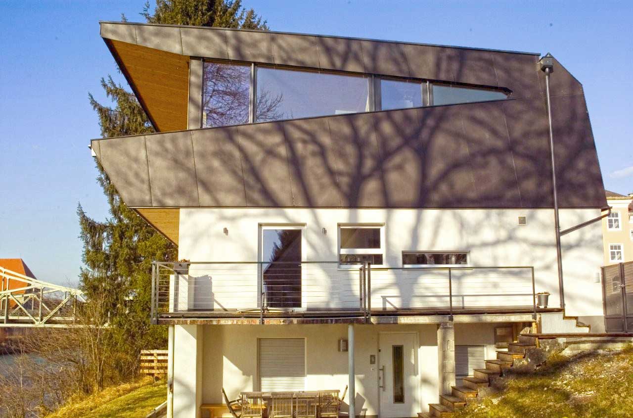 Mehrstöckiges modernes Haus mit schrägem Dach, großen Fenstern, weißem Unterbau, Balkon mit Metallgeländer und Außentreppe, gelegen an einem Hang mit Blick auf Wasser und Bäume.