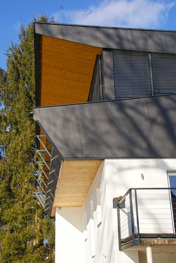 Modernes Wohnhaus mit dunklem Metalldach, Holzuntersicht und weißem Unterbau mit Balkon, Metallgeländer und Stahlseilen, umgeben von hohen Nadelbäumen unter blauem Himmel.