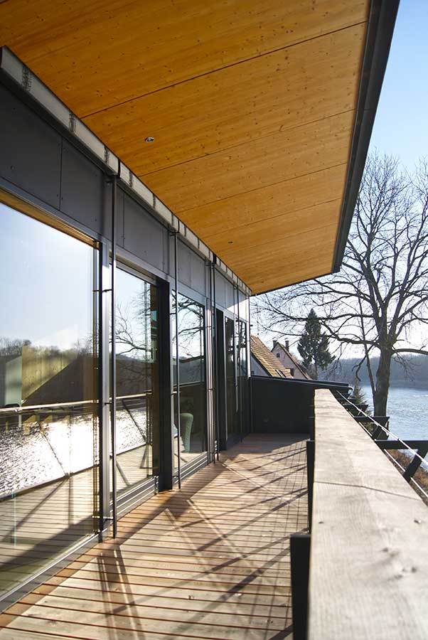 Moderner Balkon mit Holzboden, Holzdecke, großen Glasschiebetüren und Blick auf Wasser, Bäume und Dächer.