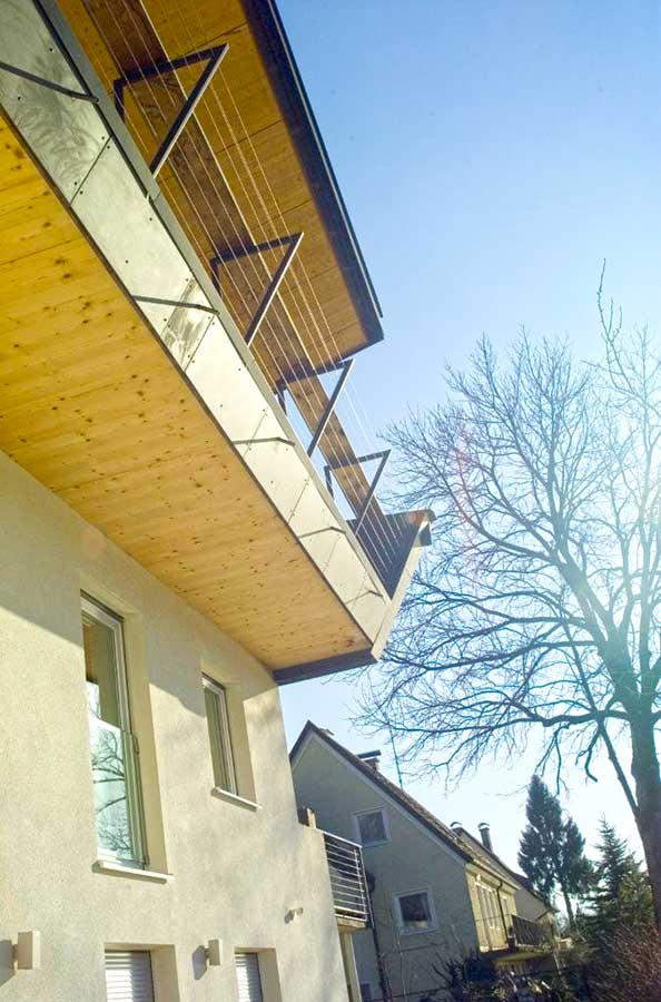 Außenansicht eines modernen Wohnhauses mit hellem Holz‑Balkon, Metallgeländer und schrägen Stützen, umgeben von Nachbarhäusern und einem kahlen Baum bei sonnigem Wetter.