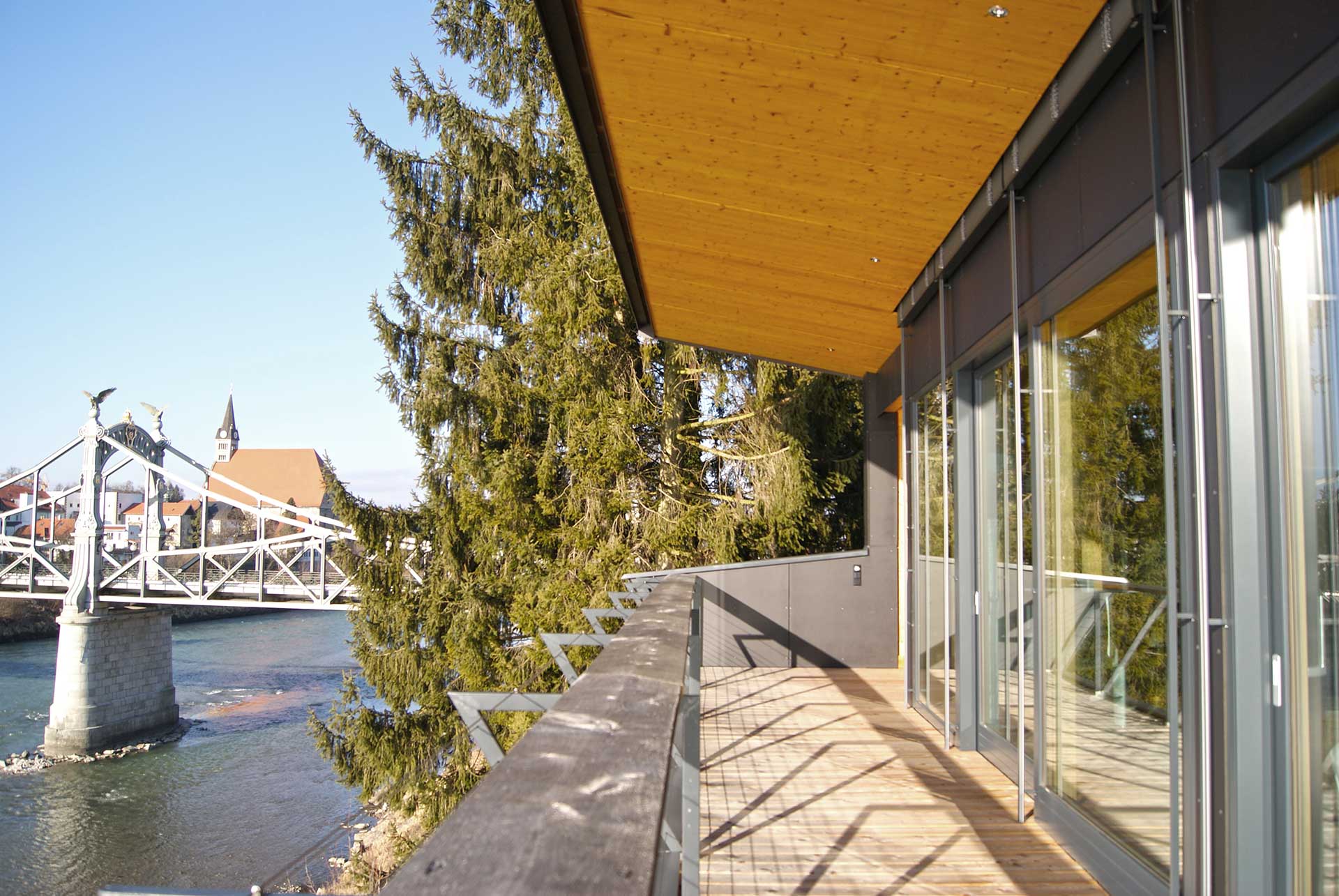 Moderner Balkon mit Holzverkleidung, großen Glasschiebetüren, Metallgeländer und Blick auf Fluss, Brücke und Altstadt mit Kirchturm.