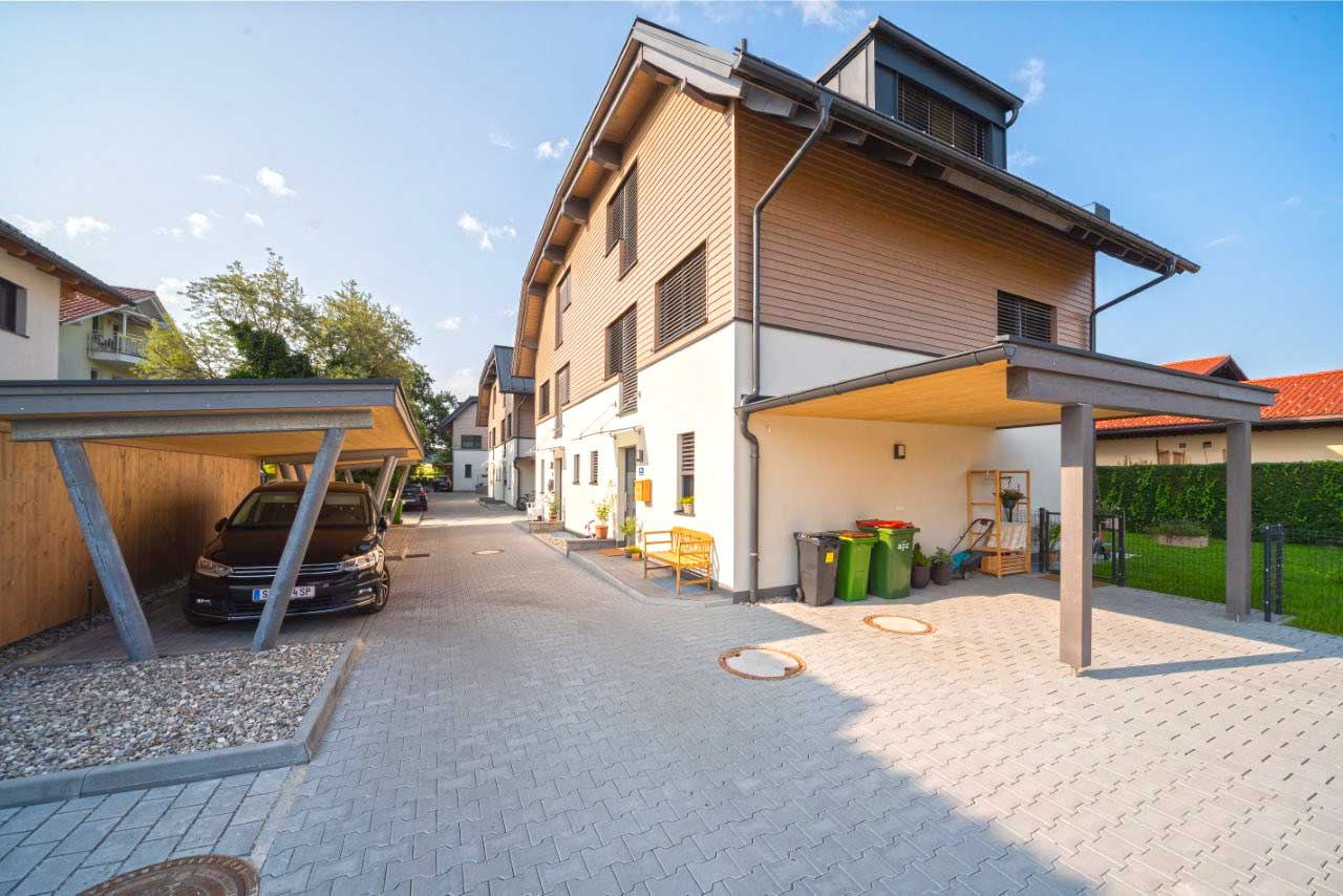 Moderne Reihenhausanlage mit schrägen Dächern, hellem Putz, Holzverkleidung, Carport mit schwarzem Auto sowie überdachtem Bereich mit Mülltonnen und Gartenutensilien.