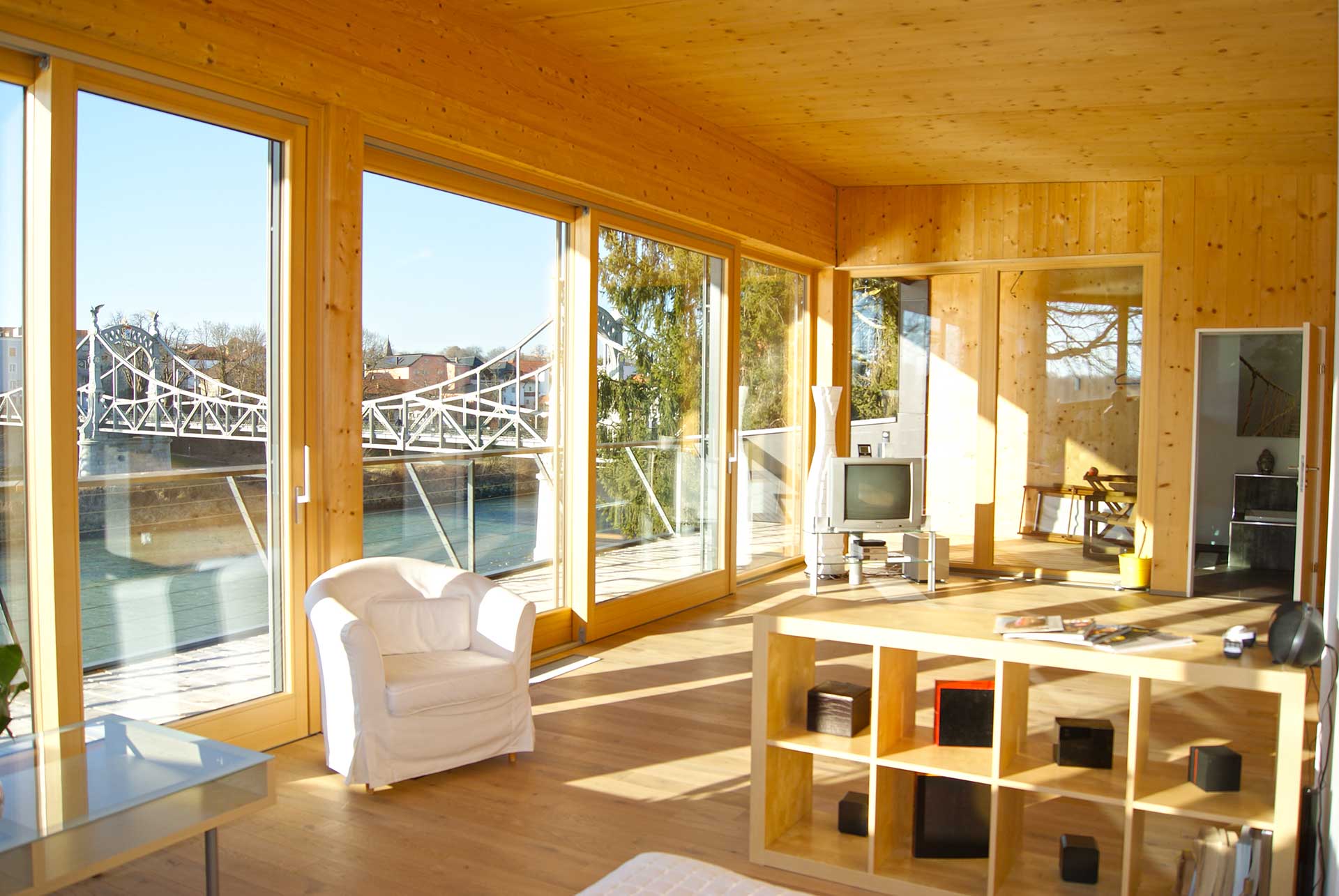 Helles Wohnzimmer mit viel hellem Holz, bodentiefen Fenstern mit Blick auf Fluss und Brücke, weißem Sessel, Holzregal und Glastisch