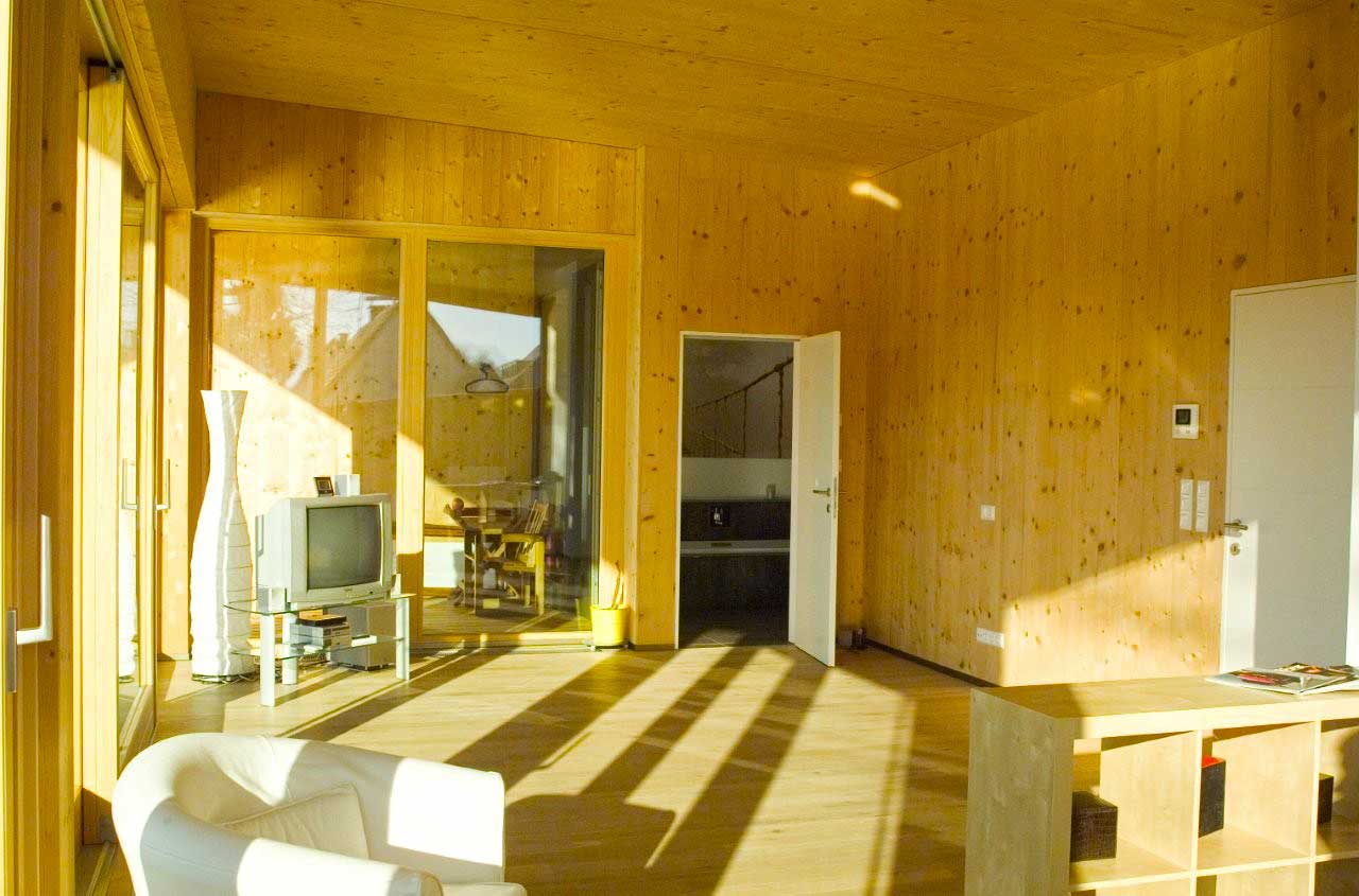 „Helles Wohnzimmer mit viel Holz an Wänden, Decke und Boden, großen Glasfenstern, weißem Sessel, TV‑Bereich und Blick in den angrenzenden Essraum.