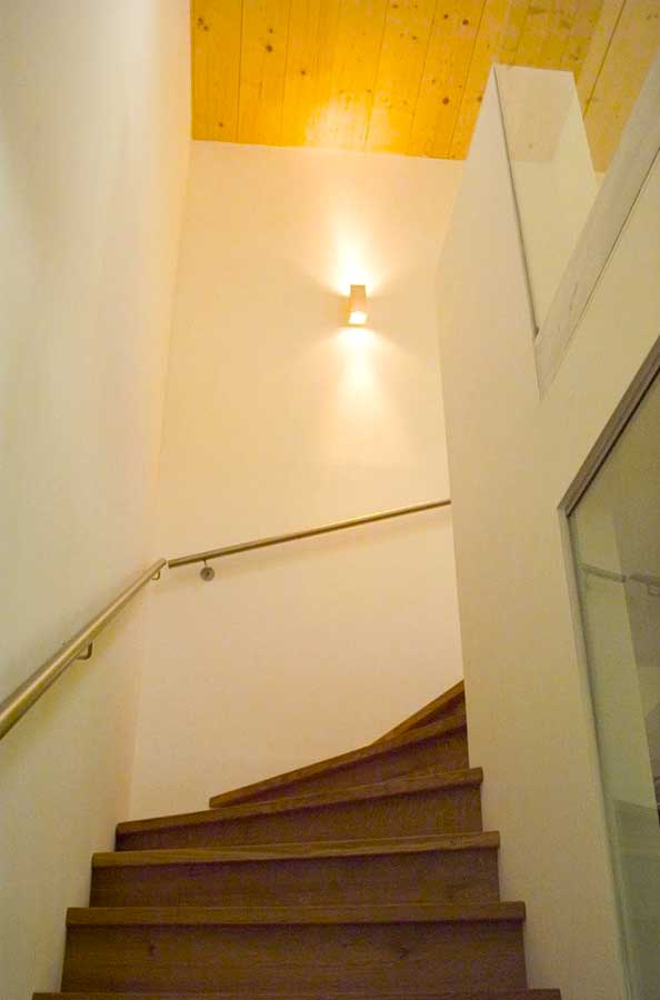 Innenraumtreppe aus Holz mit gebogener Form, weißer Wand, Metallhandlauf und warmem Wandlicht unter einer glänzenden Holzdecke.