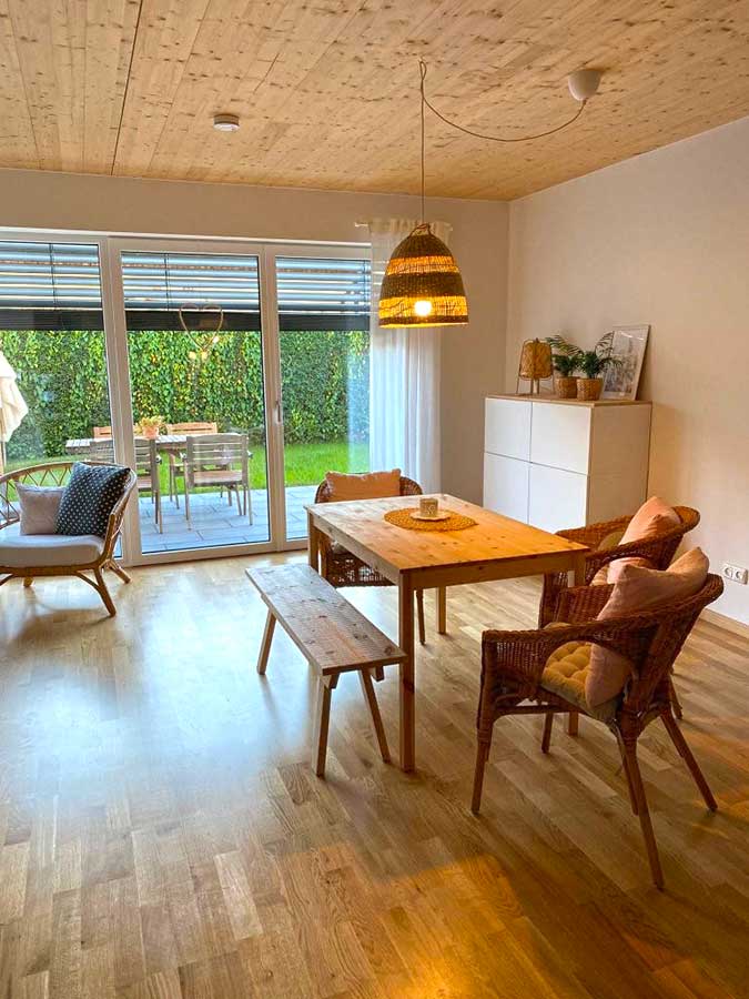 Essbereich mit Holzboden und Holzdecke, Holztisch mit Bank und Korbstühlen, Hängelampe, weißem Sideboard mit Pflanzen sowie Sessel vor großen Glasschiebetüren zum Garten.