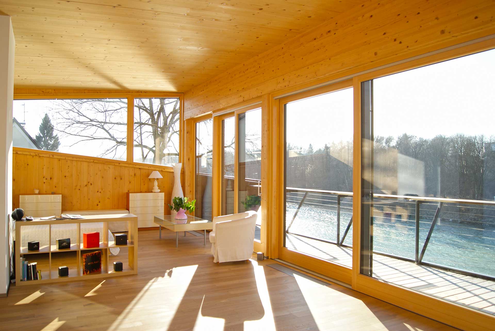 Heller Wohnraum mit viel hellem Holz, bodentiefen Fenstern zum Balkon mit Wasserblick, weißem Sessel, Couchtisch mit Vase und Regal mit Deko.