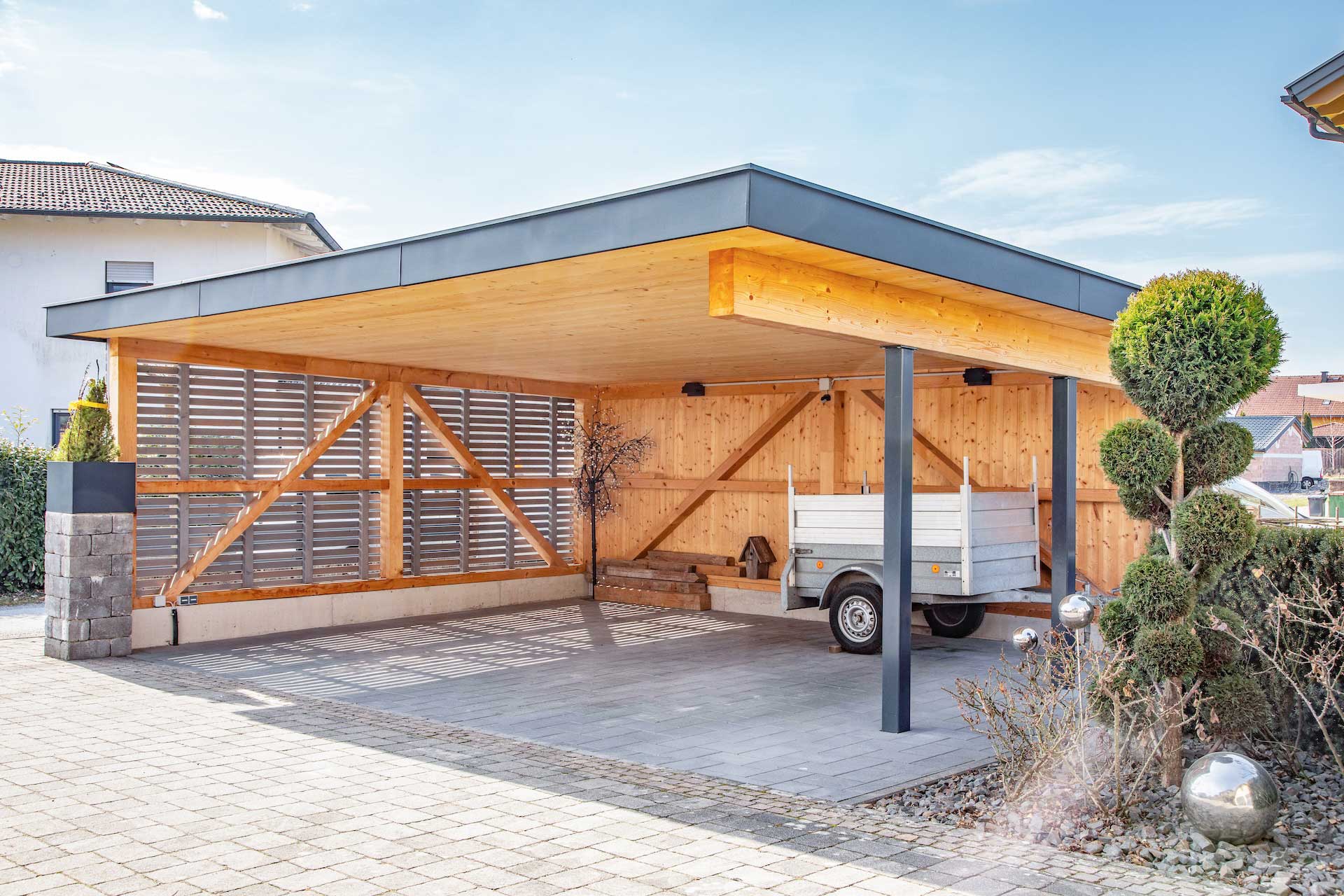 Moderner Carport mit Holzverkleidung, schwarzen Metallstützen, flachem Dach und abgestelltem Anhänger auf gepflasterter Einfahrt, umgeben von gepflegten Sträuchern. © SARA BUBNA photography