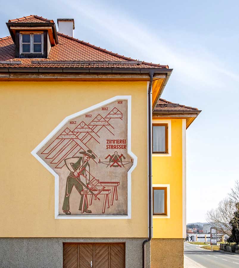 Fassadenmalerei an gelbem Gebäude, die einen Zimmerer beim Sägen an einer Werkbank zeigt, umrahmt von einem stilisierten Hausdach und dem Schriftzug Zimmerei Strasser © SARA BUBNA photography