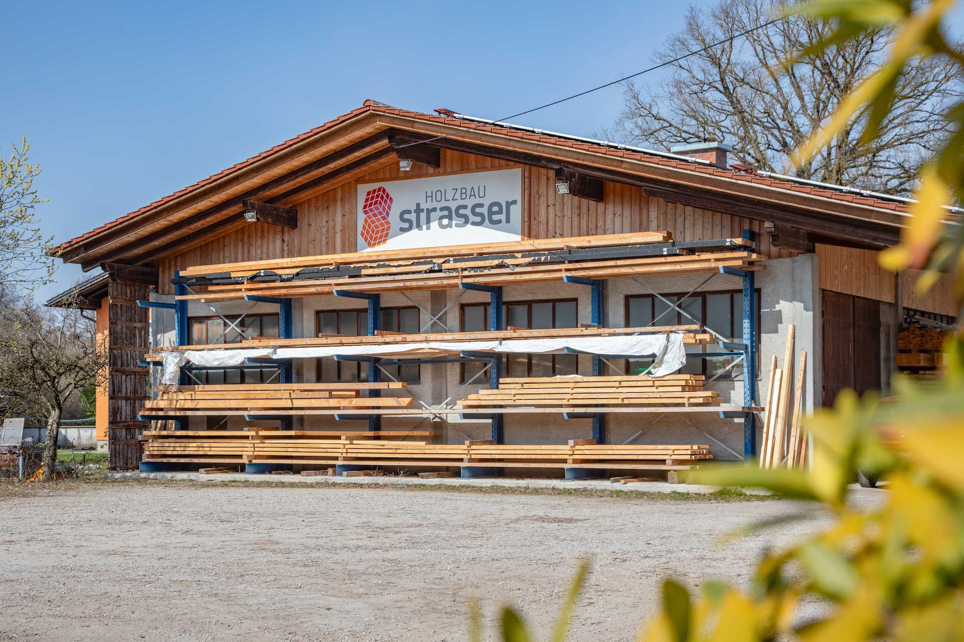 Holzbau‑Gebäude mit Satteldach und außen montierten Metallregalen, auf denen lange Holzbohlen gestapelt sind, darüber ein Schild mit der Aufschrift Holzbau Strasser © SARA BUBNA photography