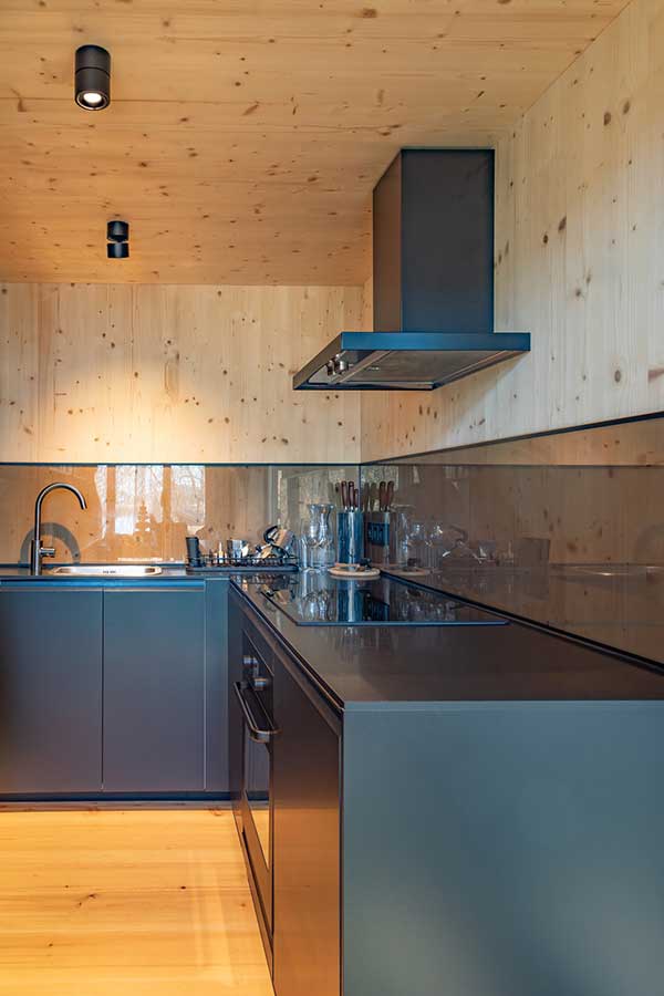 Minimalistische Küche mit hellen Holzwänden und -decke, dunklen Schränken und Arbeitsflächen, Induktionskochfeld, eingebautem Ofen, Edelstahl-Dunstabzug, Spüle mit schwarzer Armatur und ordentlich arrangierten Küchenutensilien. © SARA BUBNA photography