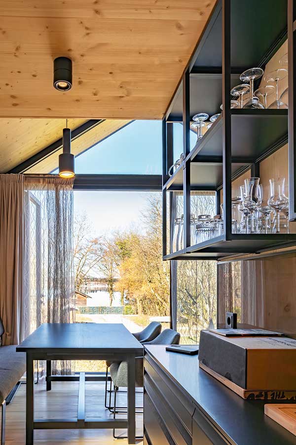 Moderner Innenraum mit hoher schräger Holzdecke, großen bodentiefen Fenstern mit Blick auf Bäume und Wasser, schwarzem Esstisch mit Stühlen, Arbeitsfläche mit Schubladen und darüber einem Regal mit Gläsern. © SARA BUBNA photography