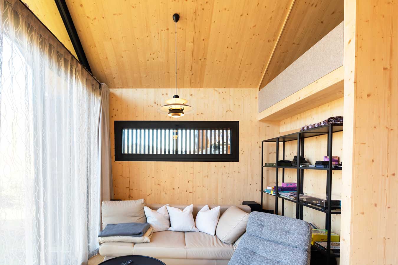 Gemütlicher moderner Wohnraum mit hellen Holzwänden und -decke, Sofa mit Kissen, rundem Couchtisch, grauem Sessel, schwarzem Metallregal mit Decken und Büchern, darüber ein Loftbereich, hängender Lampe und großem Fenster mit weißen Vorhängen. © SARA BUBNA photography