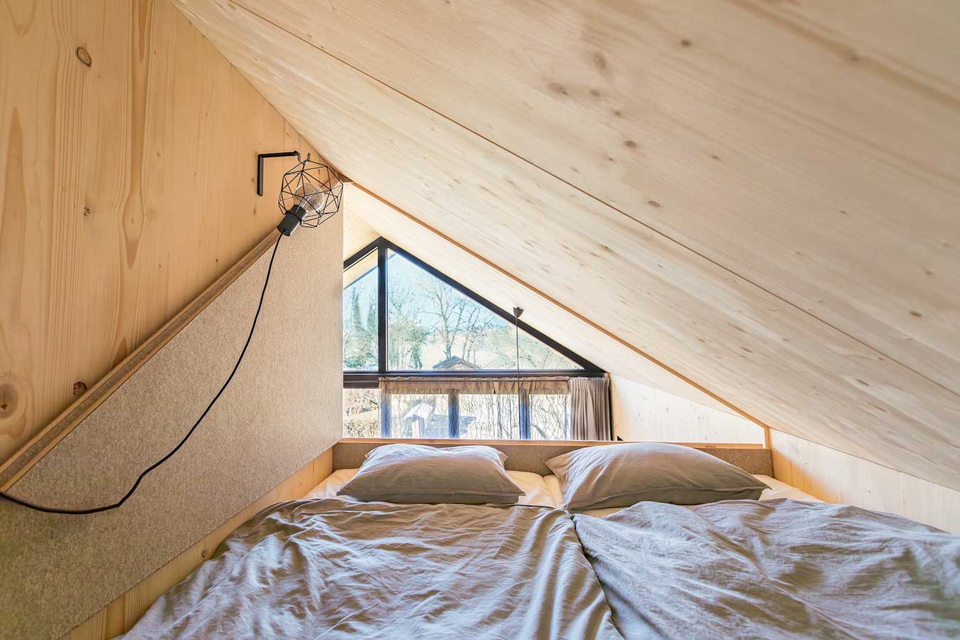 Gemütlicher Schlafbereich im Loft mit schräger Holzdecke, Bett mit grauer Bettwäsche unter einem dreieckigen Fenster und minimalistischer Wandleuchte. © SARA BUBNA photography