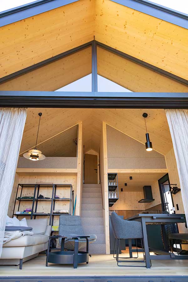 Innenraum eines kleinen modernen Holzhauses mit hoher Dachspitze, großen Glasfronten, hellem Holz, Sofa, Essbereich, kompakter Küche und Treppe zum Schlafloft. © SARA BUBNA photography