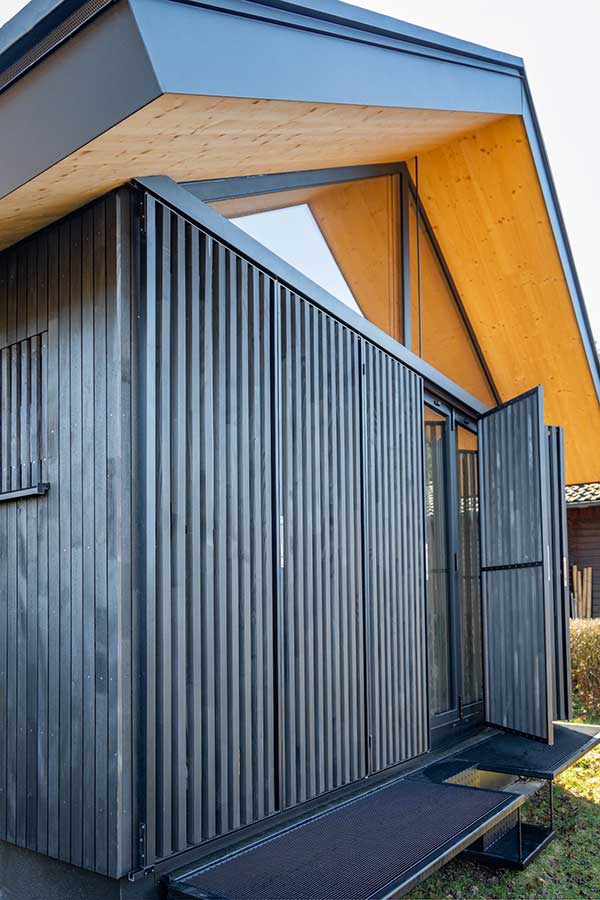 Kleines modernes Haus mit dunkler Lamellenfassade, steilem Dach mit Holzuntersicht, großer Glasfront und seitlich geöffnetem Paneel auf erhöhter Metallplattform im Garten. © SARA BUBNA photography