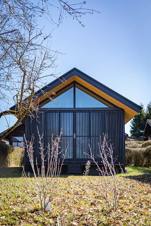 Kleines modernes Haus mit steilem Satteldach, großer dreieckiger Glasfront, dunkler Lamellenfassade und Holzuntersicht, umgeben von Rasen, Sträuchern und blauem Himmel. © SARA BUBNA photography