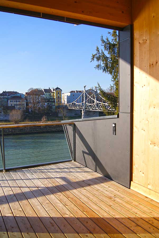 Holzterrasse mit Glas‑Metall‑Geländer und freiem Blick auf einen Fluss, eine Brücke und städtische Gebäude bei sonnigem Wetter.