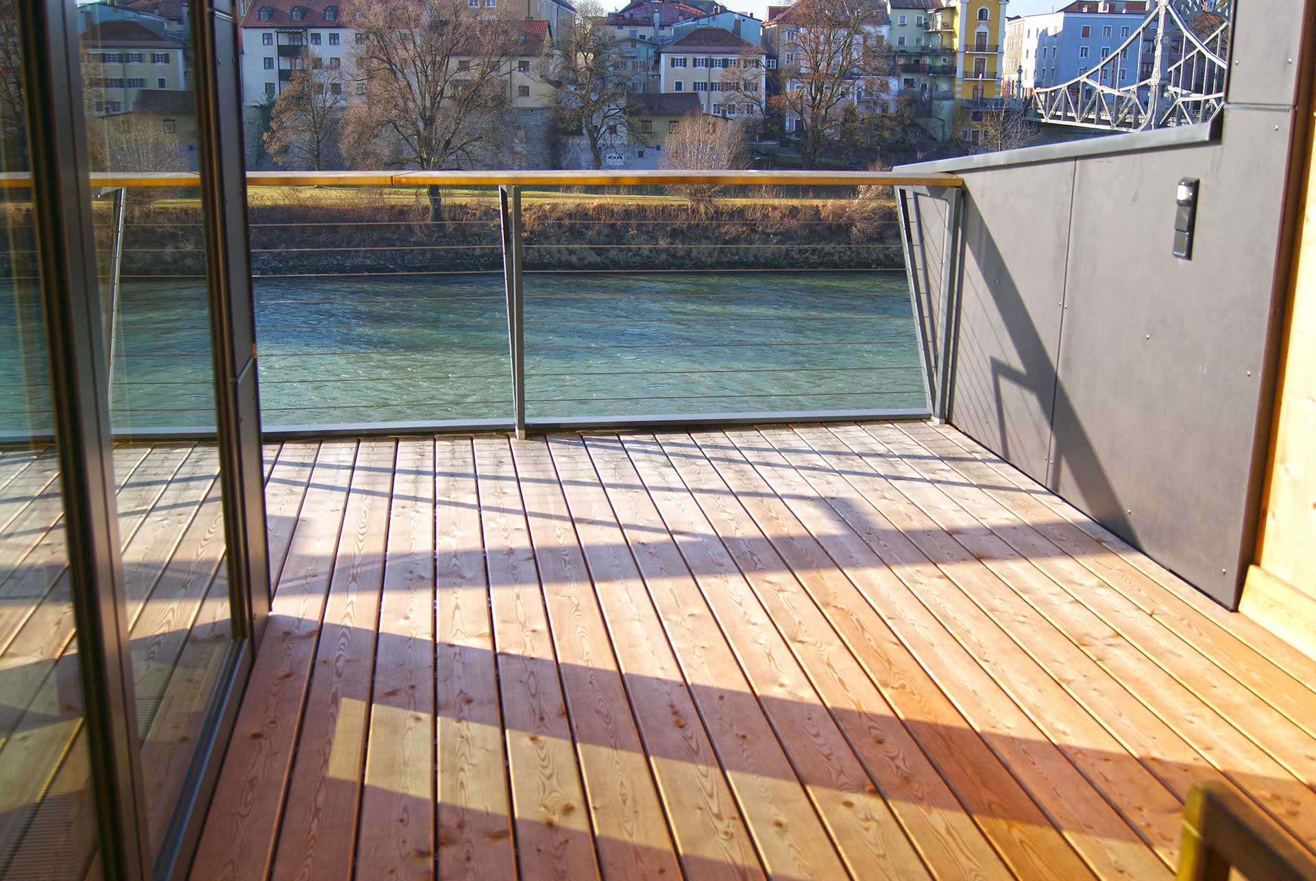 Holzterrasse mit Glas‑Metall‑Geländer und freiem Blick auf einen Fluss, eine Brücke und städtische Gebäude bei sonnigem Wetter.