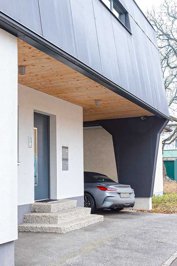 Moderne Hausfassade mit Carport, grauem Sportwagen, Holzdecke und grauer Eingangstür mit zwei Briefkästen. © SARA BUBNA photography