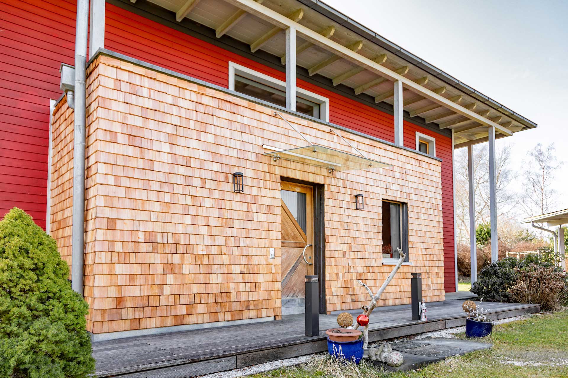 Moderner Holzanbau mit Schindelfassade vor einem roten Holzhaus, überdachtem Eingangsbereich und Terrasse.